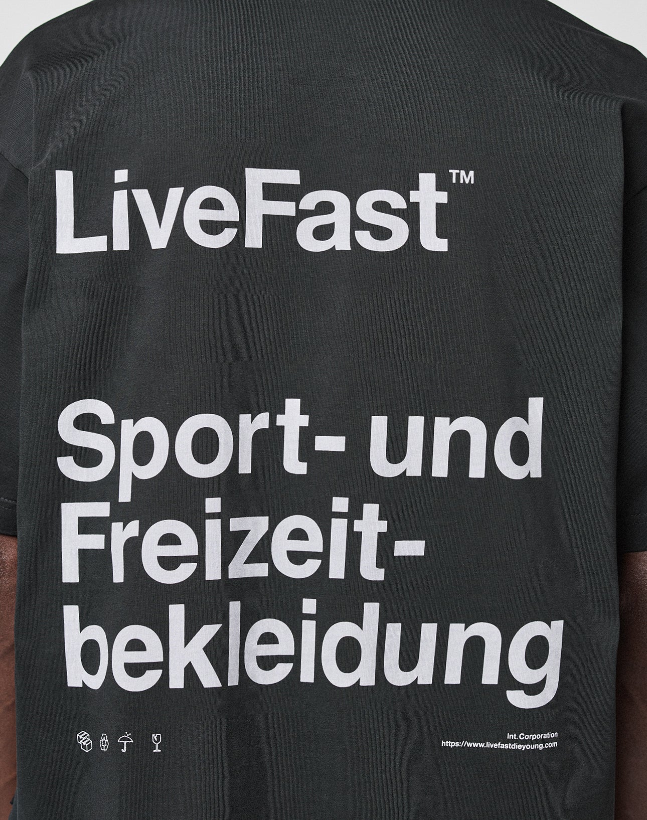 A close-up of the back of the black LIVE FAST DIE YOUNG Basic Trademark Tee shows large white text: LiveFast™ Sport- und Freizeitbekleidung. Small icons and a URL appear at the bottom. 100% Baumwolle, markanter Front- und Backprint.