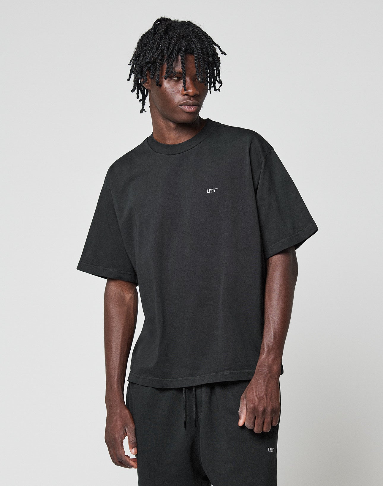 A man with short dreadlocks wears the LIVE FAST DIE YOUNG Basic Trademark Tee, a loose-fitting black T-shirt with front and back print, oversize fit, and 100% cotton, paired with black shorts against a light gray background.