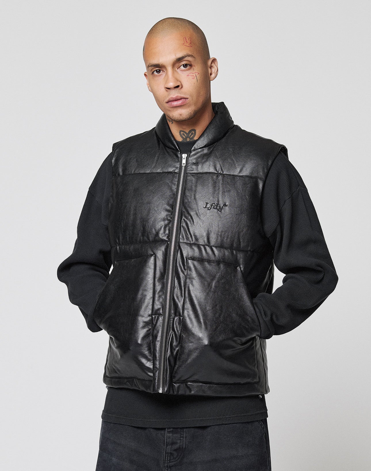 A person with a shaved head and face tattoos wears the LIVE FAST DIE YOUNG Padded Vest in black, regular fit, with padded stand-up collar over a black pullover, hands casually in pockets, standing against a plain light gray background.