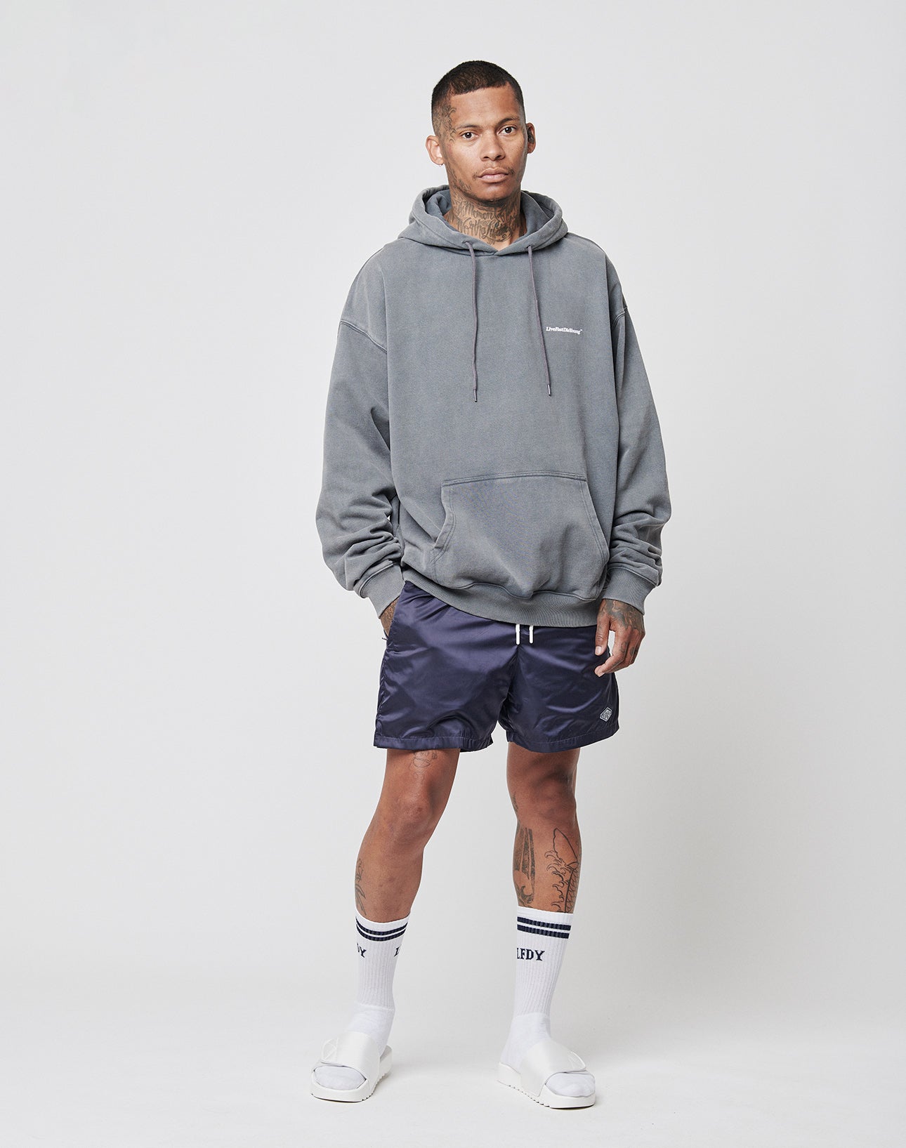 A man with short hair and visible tattoos wears a gray Basic Heavy Hooded by LIVE FAST DIE YOUNG, navy blue shorts, white crew socks with black stripes, and white slide sandals against a plain white background.