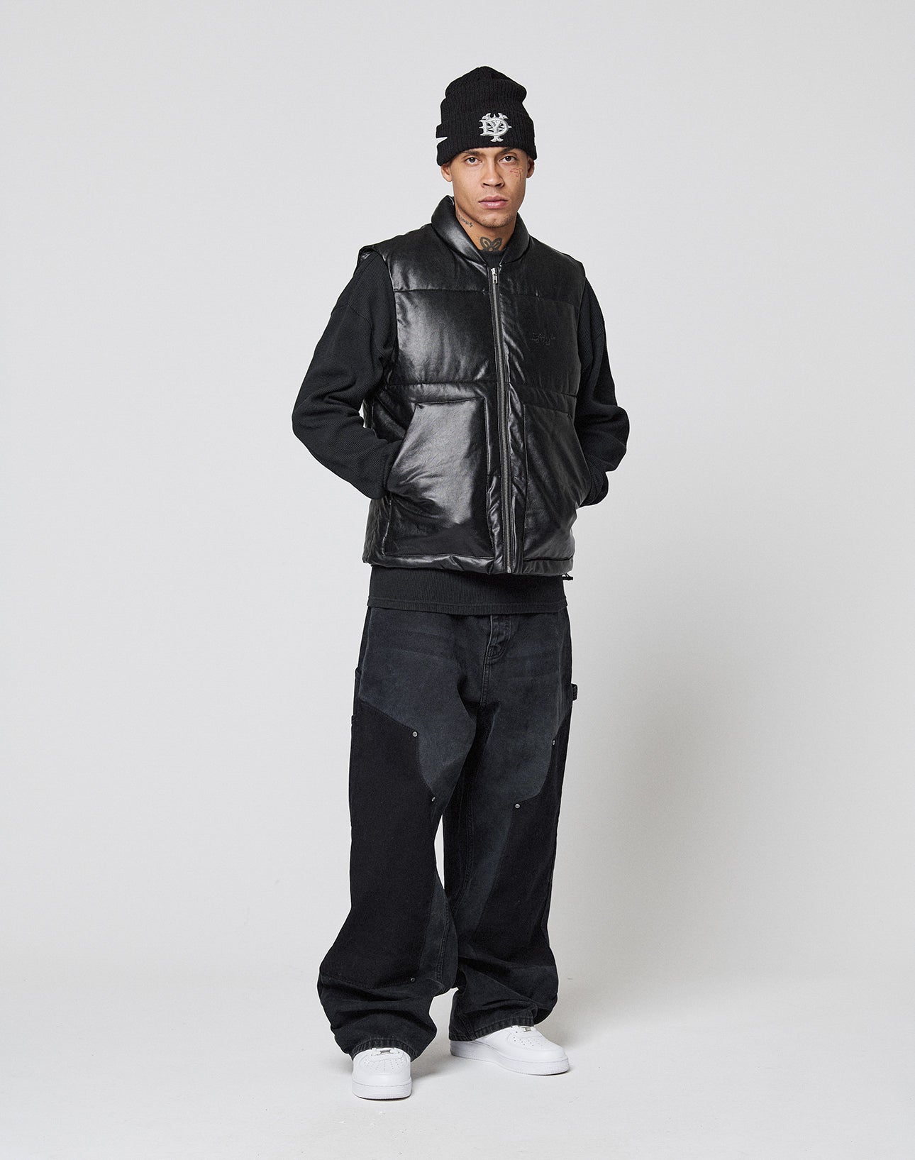 A person stands against a plain background wearing a black beanie, LIVE FAST DIE YOUNG Padded Vest with a stand collar, black long-sleeve shirt, loose regular fit black jeans, and white sneakers, looking at the camera with hands in the vest pockets.