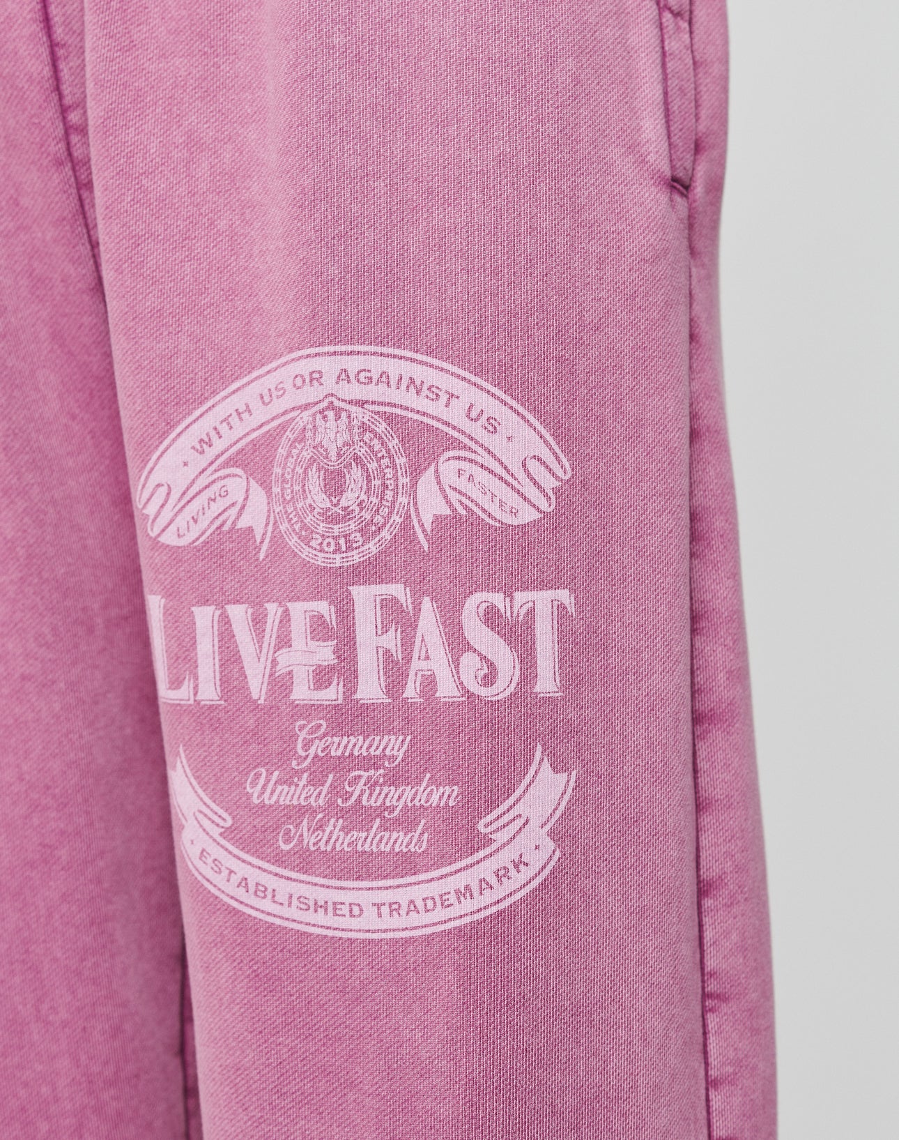 The Loyal Loose Jogger by LIVE FAST DIE YOUNG features pink Baumwolle fabric with white āLIVE FASTā text, banners, a circular emblem, and country names. Oversize fit and vintage wash deliver a bold look.