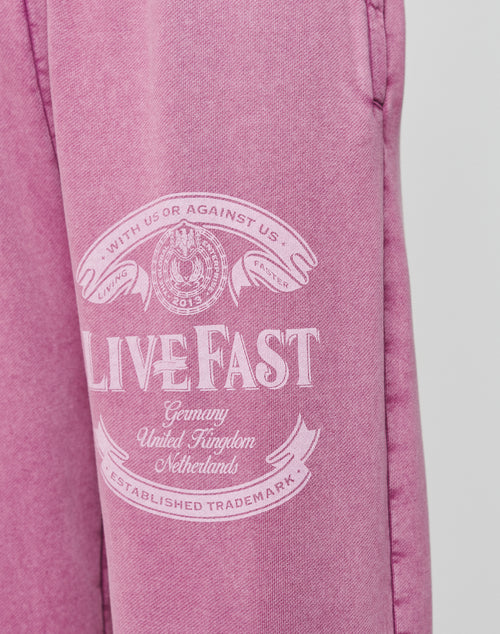 The Loyal Loose Jogger by LIVE FAST DIE YOUNG features pink Baumwolle fabric with white “LIVE FAST” text, banners, a circular emblem, and country names. Oversize fit and vintage wash deliver a bold look.