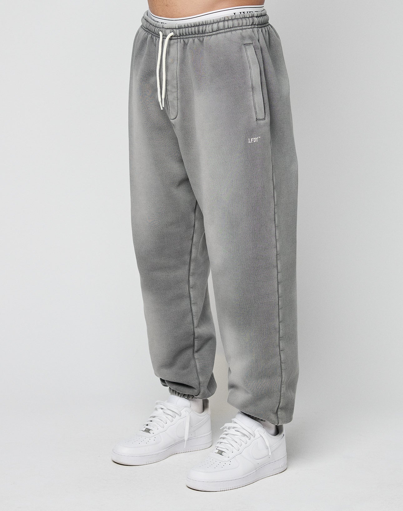 A person models the LIVE FAST DIE YOUNG Basic Loose Joggerāgray, loose-fitting with a faded look, drawstring waist, and side pocketsāpaired with white sneakers, standing against a plain light background.