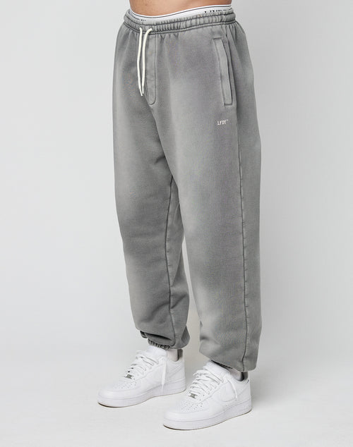 A person models the LIVE FAST DIE YOUNG Basic Loose Jogger—gray, loose-fitting with a faded look, drawstring waist, and side pockets—paired with white sneakers, standing against a plain light background.