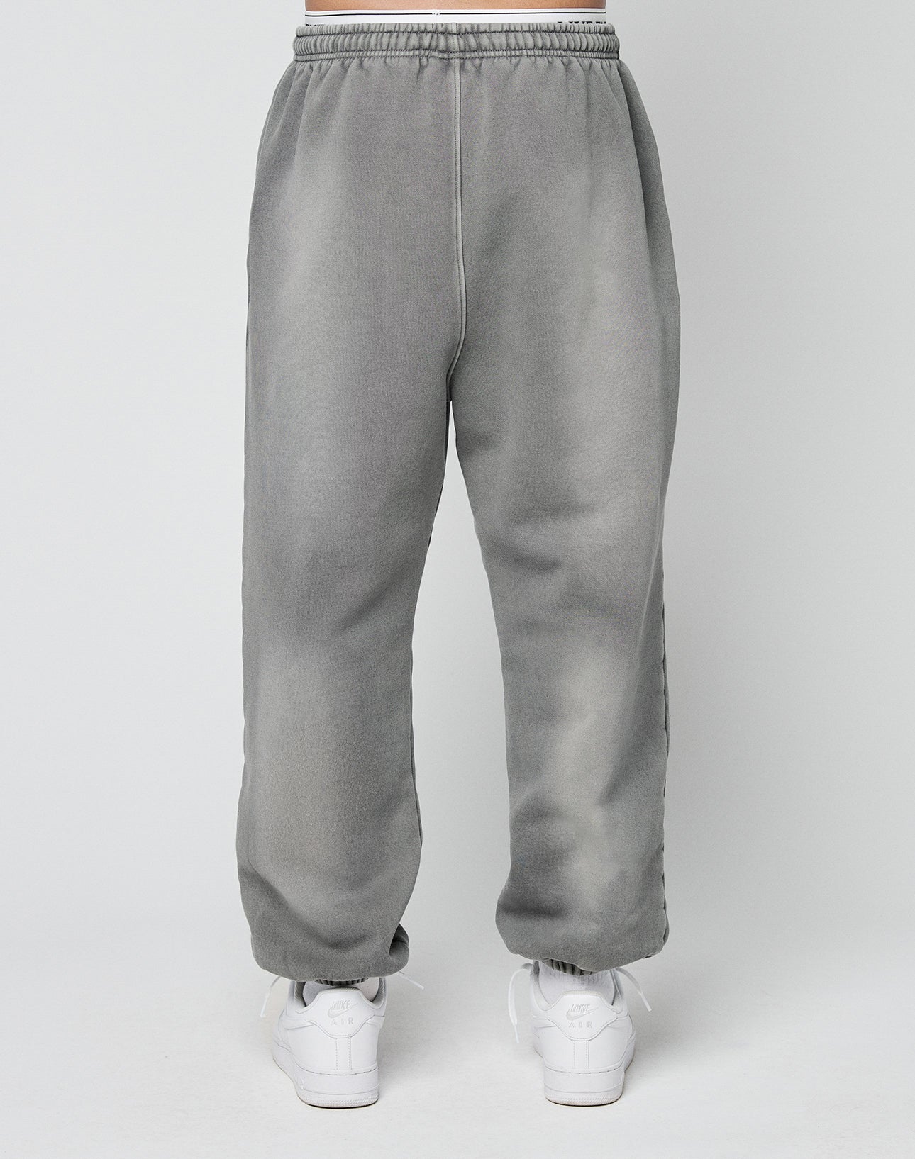 A person models the LIVE FAST DIE YOUNG Basic Loose Joggerāgray sweatpants with an elastic waistbandāpaired with white athletic shoes, standing against a plain light background.
