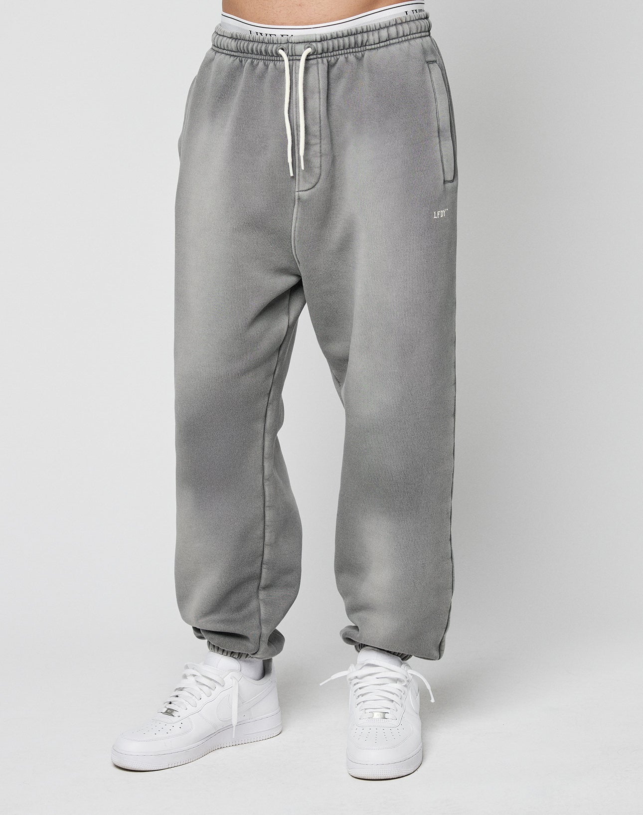 A person models the LIVE FAST DIE YOUNG Basic Loose Joggerālight gray, loose-fitting sweatpants with a logo print and white drawstringsāpaired with white sneakers against a plain light gray background.