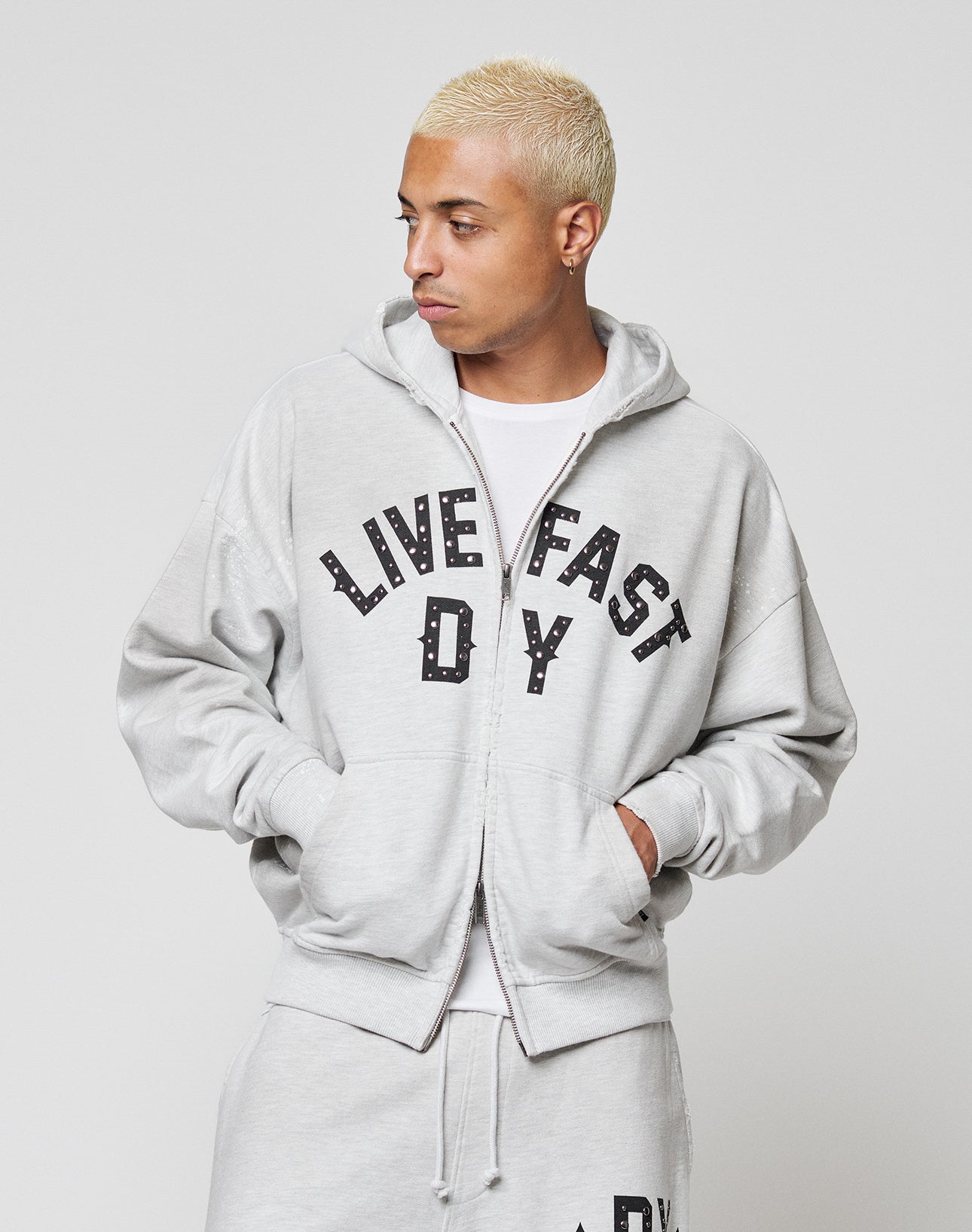 A person wearing the Live Fast Rivet Hooded Zip by LIVE FAST DIE YOUNG in light gray with bold black LIVE FAST D*Y print and subtle distressing, hands in pockets, looking to the side.