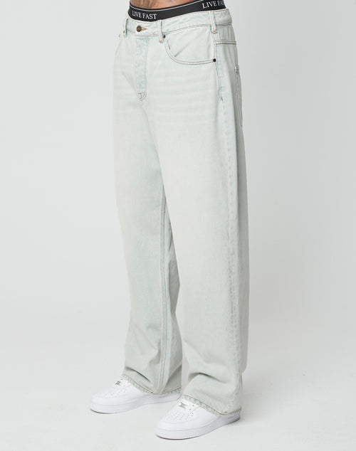 A person in LIVE FAST DIE YOUNGs LFDY Baggy Denim light-wash jeans, white sneakers, and a visible black LIVE FAST waistband stands against a plain white background.