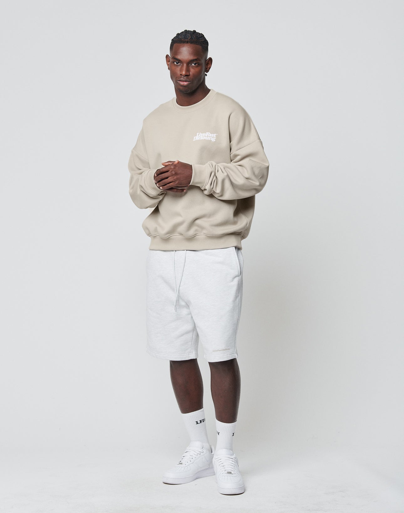A man stands against a white background, wearing the LIVE FAST DIE YOUNG Basic Chest Sweater in an oversize beige fit, light gray shorts, white crew socks, and white sneakers. He faces forward with his hands clasped in front.