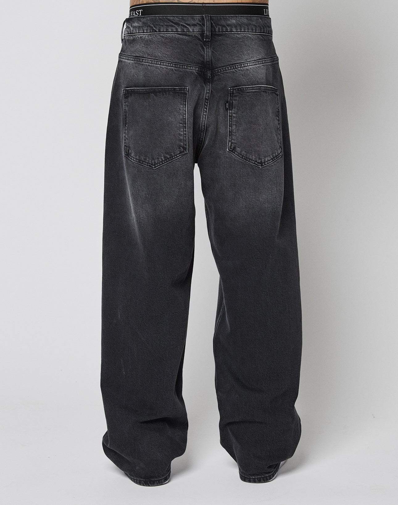 Someone wears LIVE FAST DIE YOUNG’s LFDY Tire Tracks Denim—loose-fitting, baggy black jeans with a slightly faded wash and two large back pockets—crafted from 100% Baumwolle, shown from the back against a plain light background.
