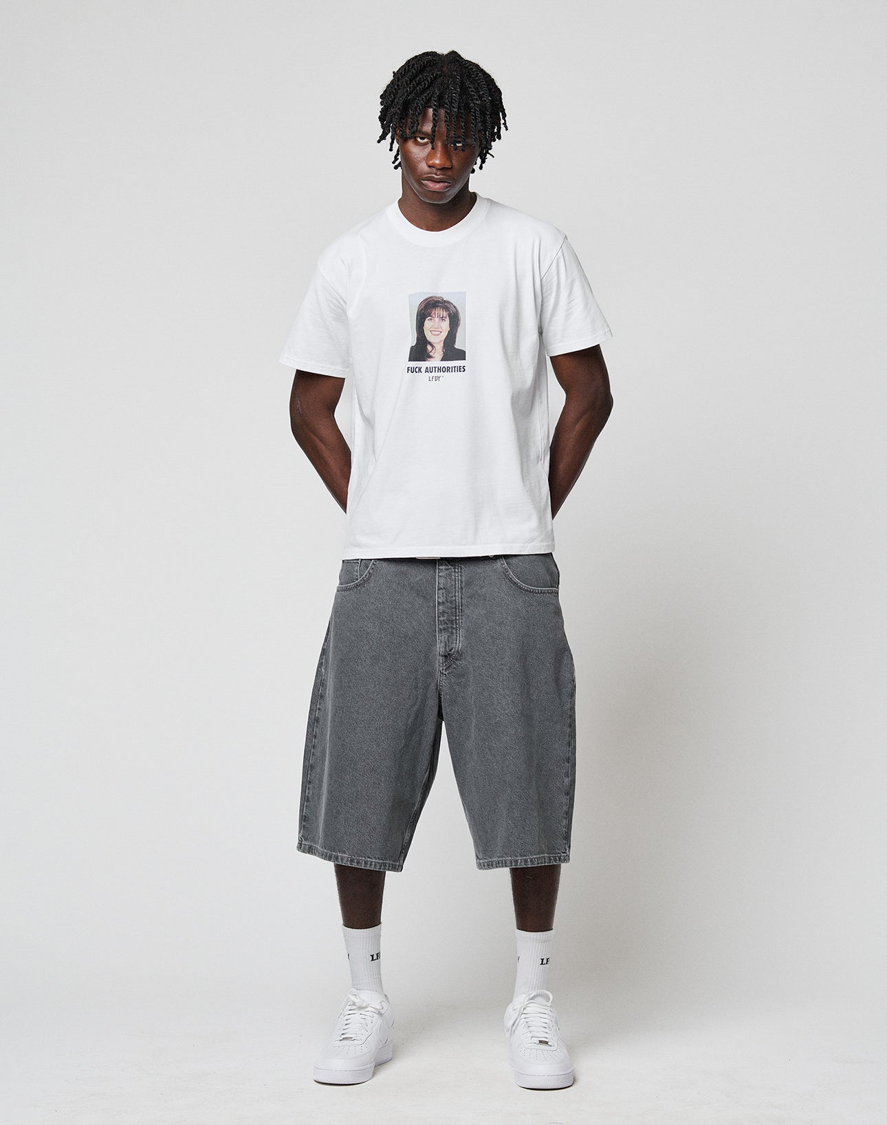 A man with short dreadlocks wears a white graphic T-shirt, LIVE FAST DIE YOUNG LFDY Jorts in a Baggy Fit, white socks, and white sneakers, standing against a plain light background.