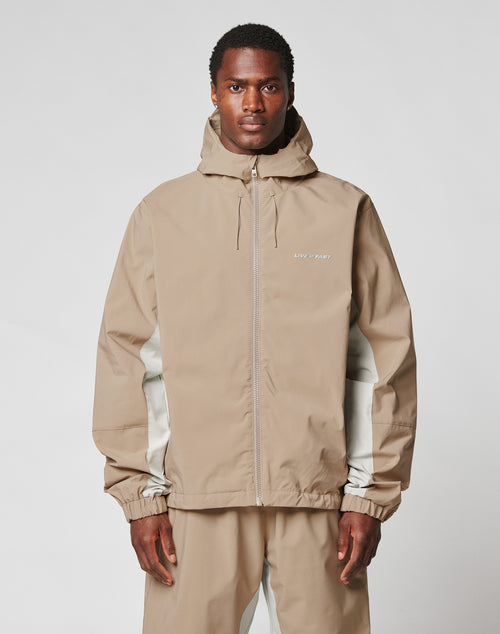 A man wears the LIVE FAST DIE YOUNG Basic Shell Jacket in light brown, featuring an oversize fit with matching pants, as he stands facing forward against a plain gray background with a neutral expression.