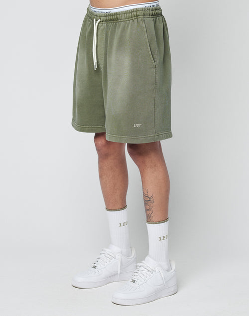 A person wears LIVE FAST DIE YOUNG Basic Sweat Shorts in olive green with an elastic waistband, paired with white LEFIT socks and white sneakers, standing against a plain light gray background. Only the lower torso and legs are visible.