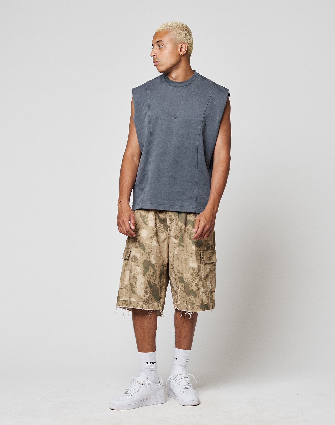 A man with short blond hair wears a LIVE FAST DIE YOUNG Heavy Muscle Tank in gray, loose camouflage cargo shorts, white socks, and white sneakers. He stands against a plain light gray background, looking to his left.