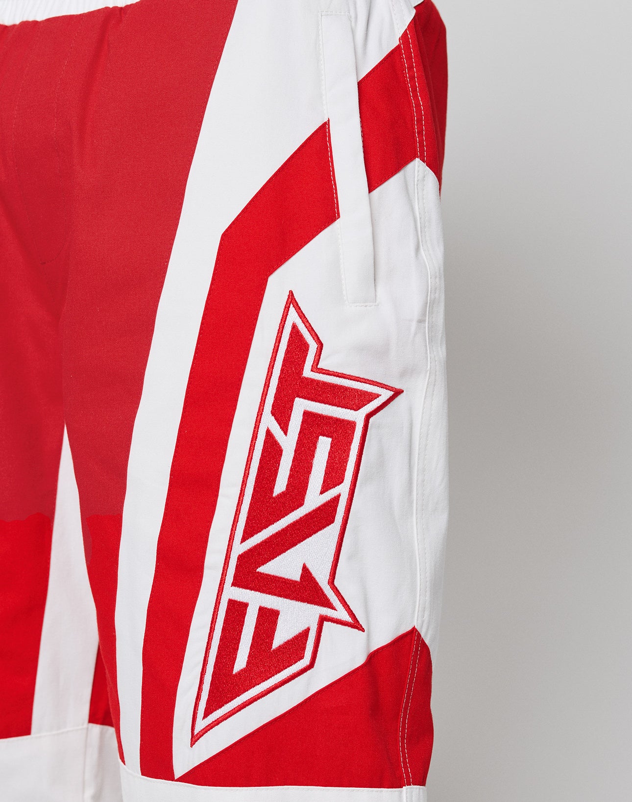 The LFDY Cross Pants by LIVE FAST DIE YOUNG are red and white oversize shorts made from 100% cotton, featuring a bold geometric pattern and an embroidered EVE patch near the pocket.