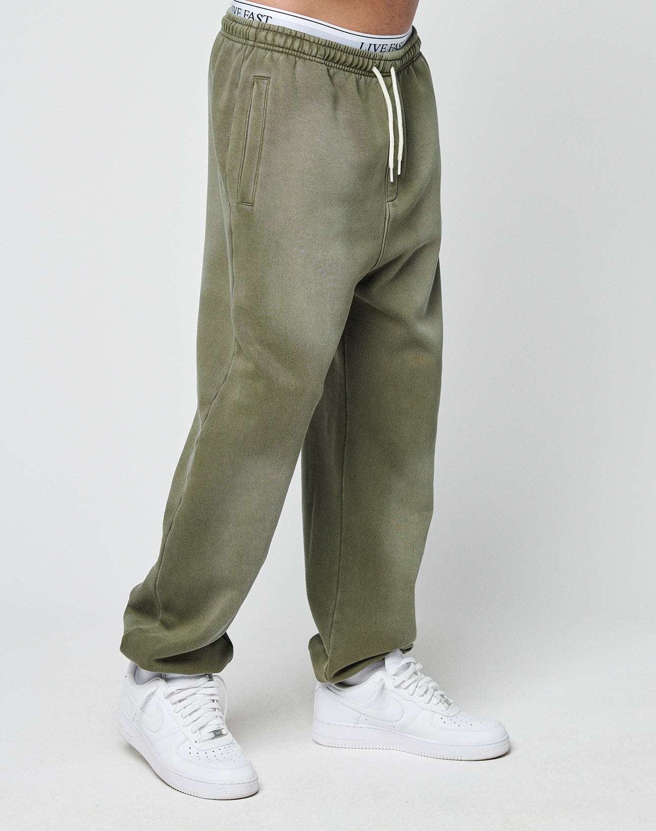 The lower half of a person is shown wearing LIVE FAST DIE YOUNG Basic Loose Jogger sweatpants in olive green with white drawstrings and white sneakers, standing against a plain light background.