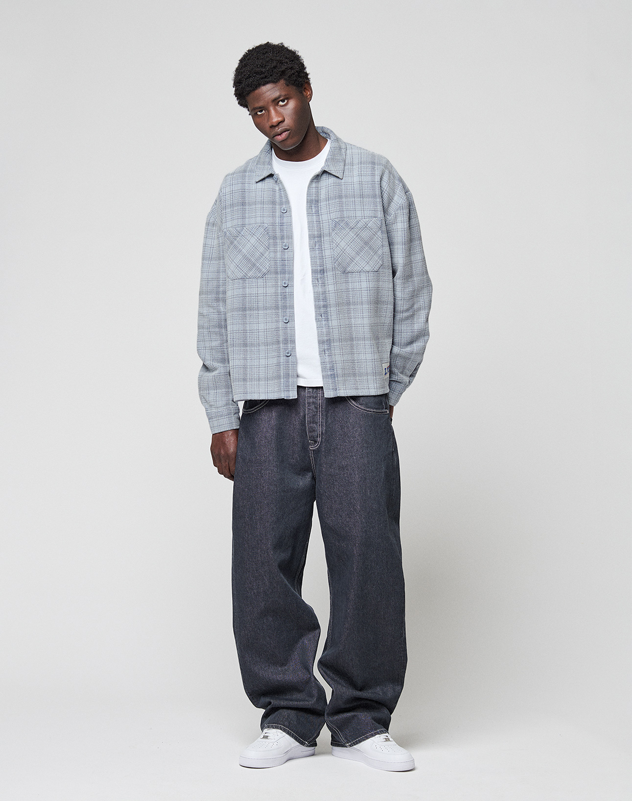 A person stands against a plain background wearing a light gray LIVE FAST DIE YOUNG Vintage Flannel over a white t-shirt, loose dark jeans in an oversize fit, and white sneakers, with a relaxed expression and posture.