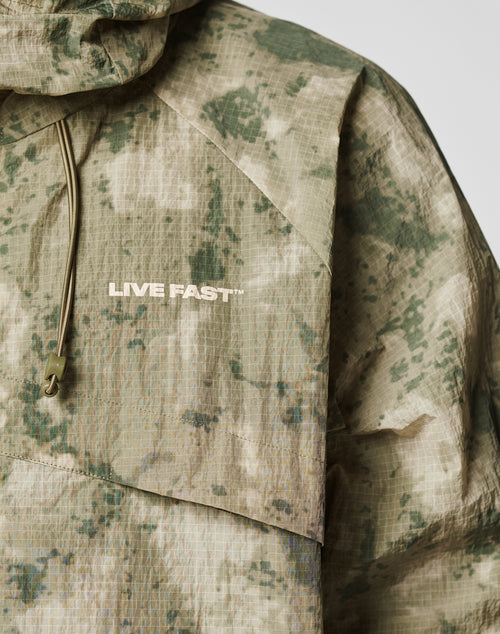 A close-up of the LIVE FAST DIE YOUNG Live Fast Nylon Jacket, featuring green and beige digital camouflage, an oversize fit, elastic hood drawstring, and “LIVE FAST™” printed in white on the chest.
