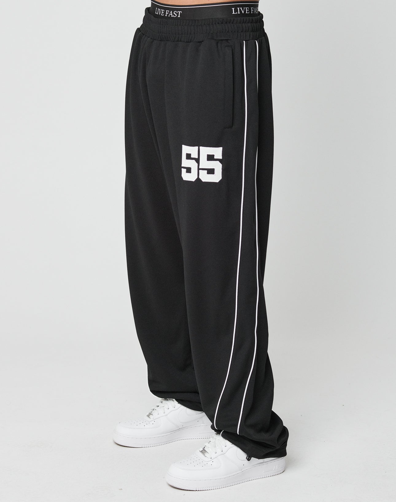 A person wears LFDY 55 Trackpants by LIVE FAST DIE YOUNG—wide fit, black with white piping, 55 printed on the left thigh, and an elastic waistband reading LIVE FAST—paired with white sneakers against a plain white background.