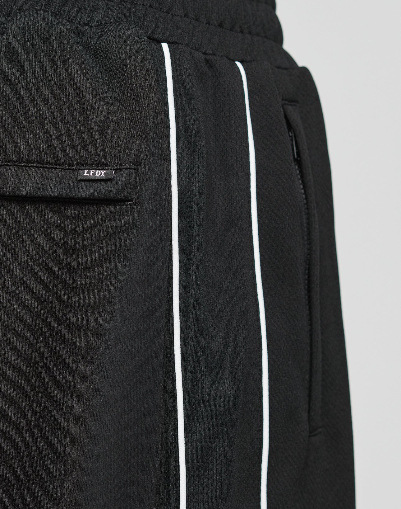 Close-up of LFDY 55 Trackpants by LIVE FAST DIE YOUNG: black wide-fit pants with white vertical stripes, zippered side pocket, elastic waistband, and LFDY tag. Crafted from soft polyester for comfort and style.