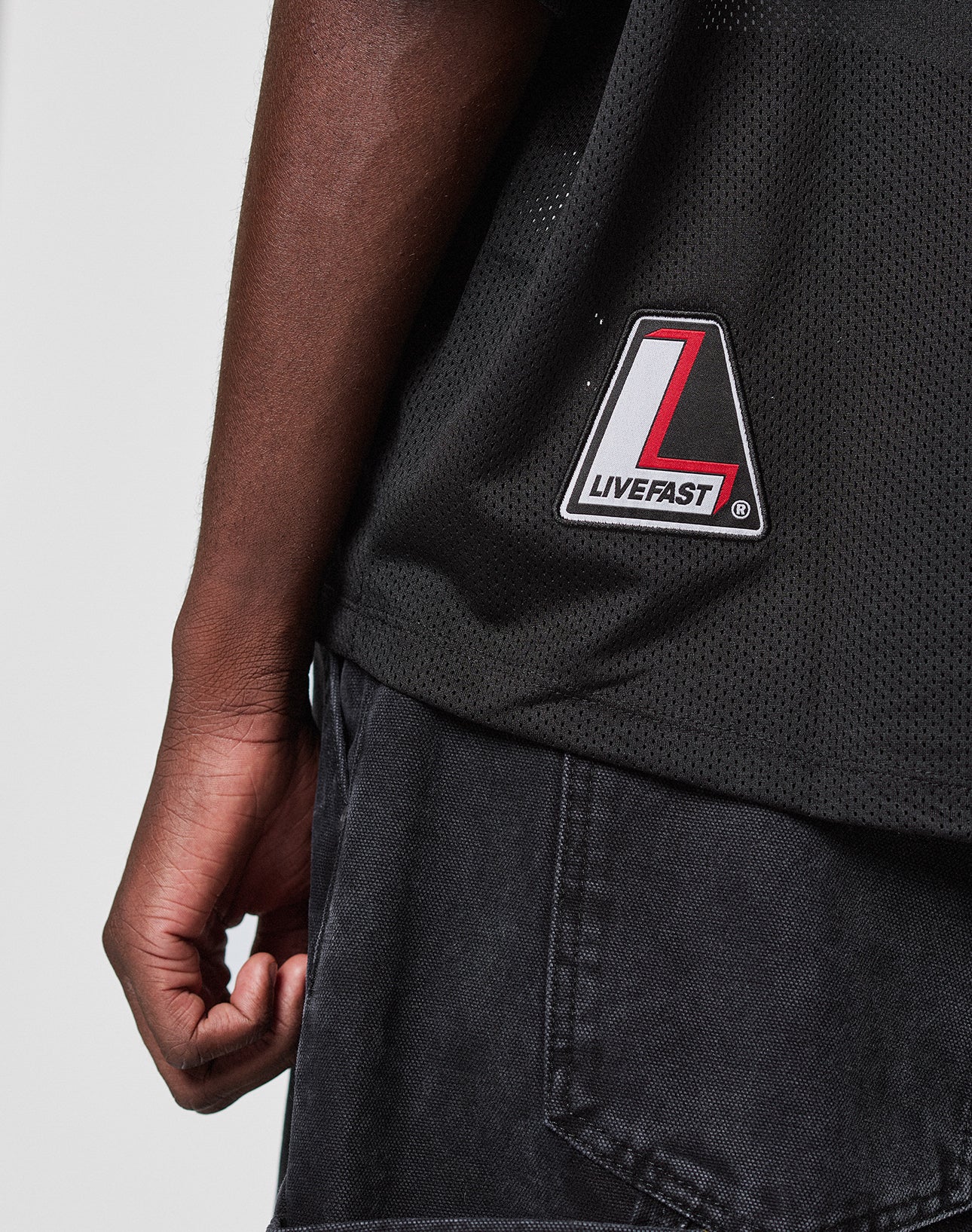 Close-up of a person's left arm and torso wearing the LFDY Mesh Jersey by LIVE FAST DIE YOUNG, featuring an oversize fit, black mesh fabric, and a striking red, white, and black patch. Styled with black denim shorts for a relaxed, on-trend look.
