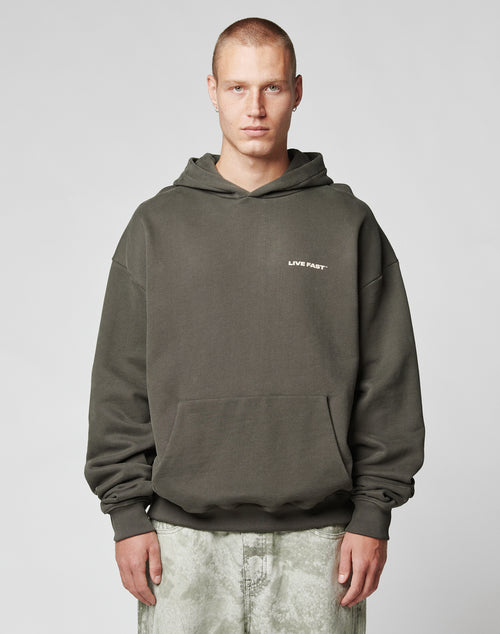 A person with short hair wears the LIVE FAST DIE YOUNG Live Fast Plain Hooded in dark olive, oversized fit with LIVE FAST on the chest, made from a soft cotton-polyester blend, paired with light marble pants against a gray background.