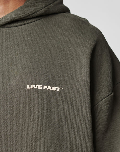Close-up of a person wearing the LIVE FAST DIE YOUNG Live Fast Plain Hooded in olive green, featuring an oversize fit and white LIVE FAST lettering on the chest. The background is plain; the persons face is not visible.