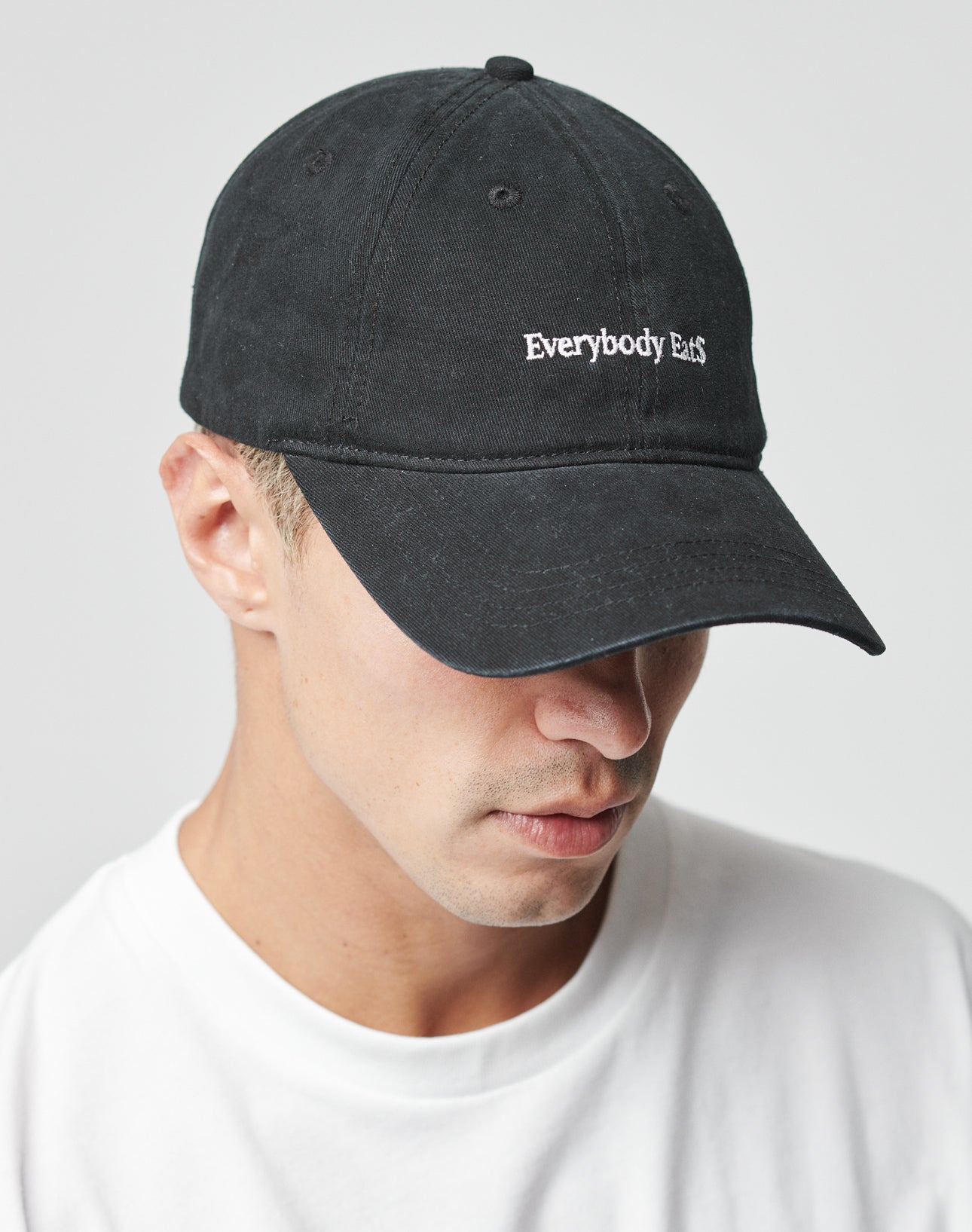 A person wearing a black LIVE FAST DIE YOUNG Everybody Eats Cap (One Size, Baumwolle, Klettverschluss, embroidered front text) and a white shirt, looking downward.
