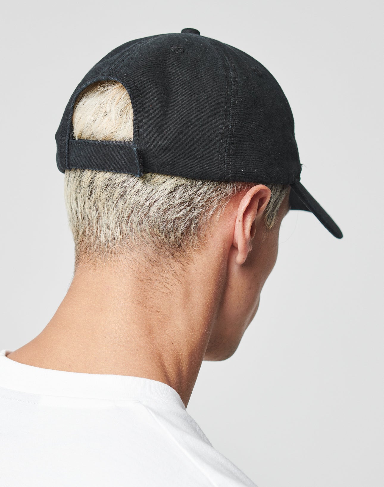 A person with short, bleached blond hair is seen from behind wearing a black LIVE FAST DIE YOUNG Everybody Eats Cap and a white shirt against a plain, light background.