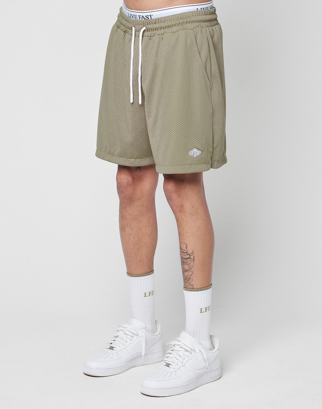 A personâs lower body is shown wearing LIVE FAST DIE YOUNG Basic Mesh Shorts in olive green polyester mesh, with an elastic waistband, drawstring, and a logo patch near the hem. They also wear white âLIVE FASTâ socks and white sneakers.