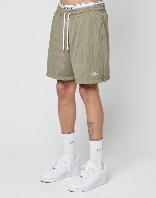 A person’s lower body is shown wearing LIVE FAST DIE YOUNG Basic Mesh Shorts in olive green polyester mesh, with an elastic waistband, drawstring, and a logo patch near the hem. They also wear white “LIVE FAST” socks and white sneakers.