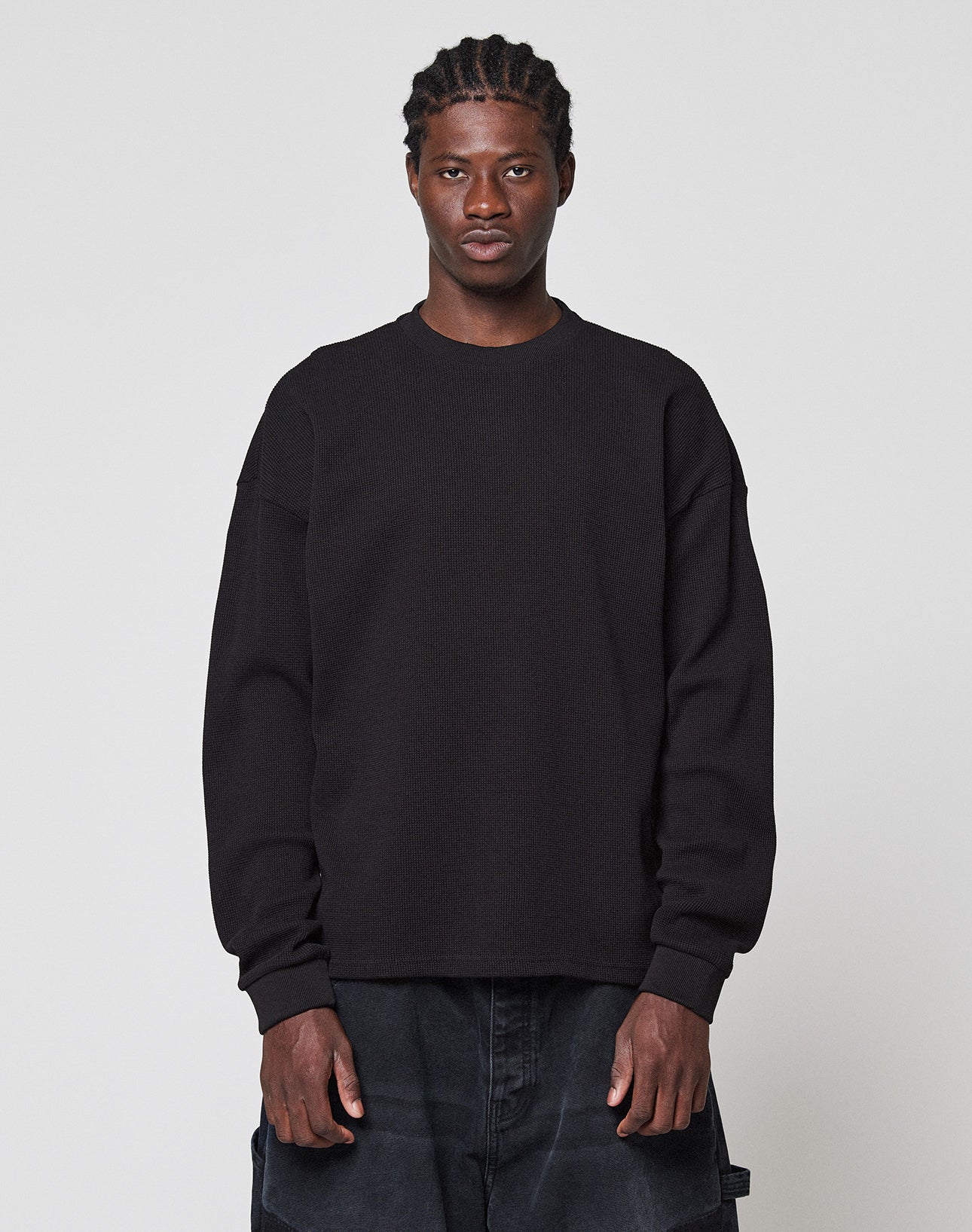 A person faces forward against a plain light gray background, wearing the LIVE FAST DIE YOUNG NY-FW Waffle Longsleeve in black with an Oversize Fit and dark pants. They have short braided hair and a neutral expression.