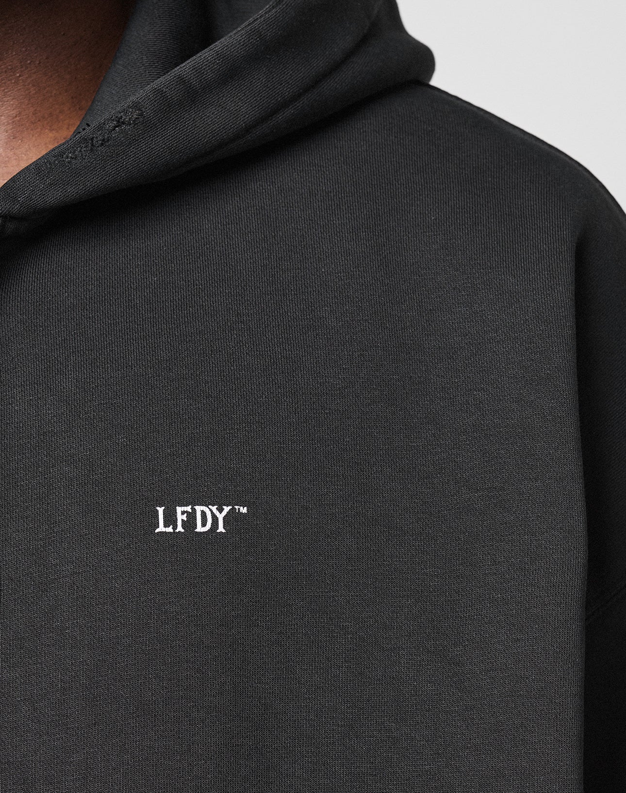 Close-up of a black LIVE FAST DIE YOUNG Basic Hooded Zip with hood up, featuring small white embroidered LFDY⢠text on the chest and subtle distressed details. Only part of the wearerās face and shoulder are visible.