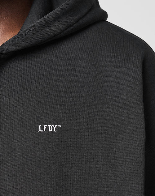 Close-up of a black LIVE FAST DIE YOUNG Basic Hooded Zip with hood up, featuring small white embroidered LFDY™ text on the chest and subtle distressed details. Only part of the wearer’s face and shoulder are visible.