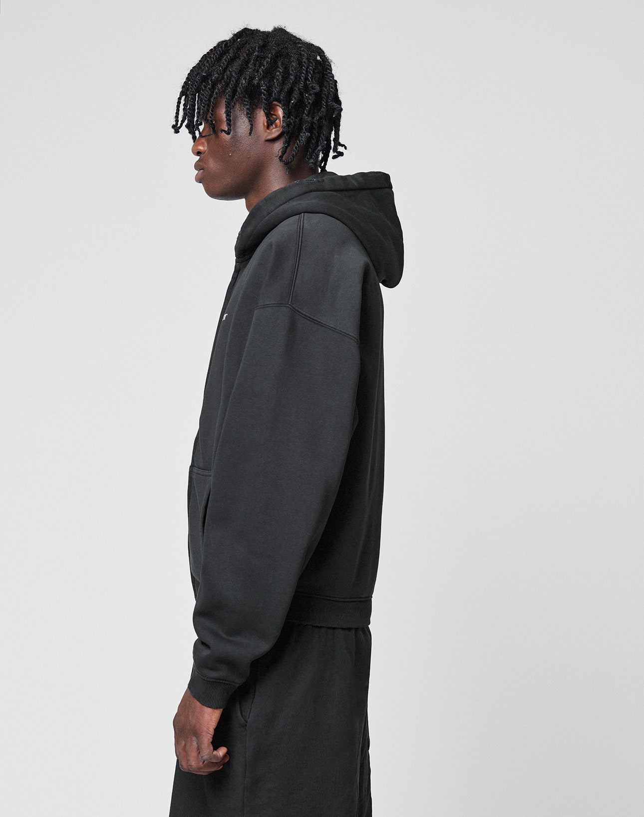 A person with short, twisted hair wears a black LIVE FAST DIE YOUNG Basic Hooded Zip and black pants, standing sideways against a plain light gray background.