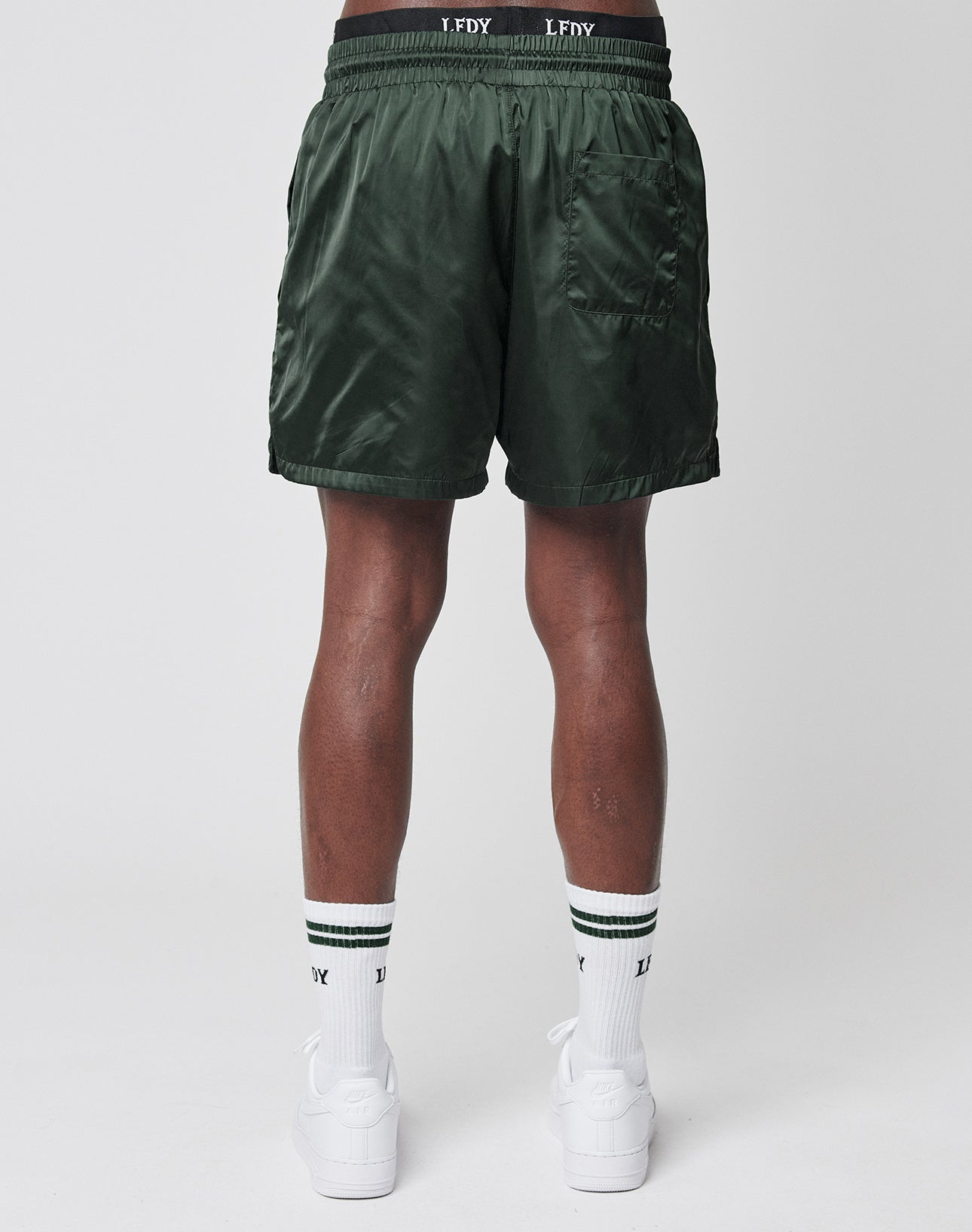 A person faces away, wearing LIVE FAST DIE YOUNG’s Basic Satin Shorts in dark green with an elastic waistband featuring repeated “LFDY” branding. Made from polyester, the shorts are styled with white socks with black stripes and white sneakers.