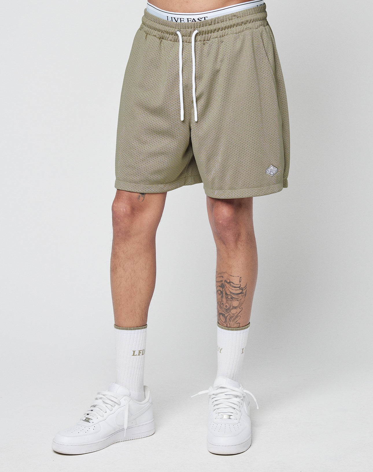A person wears LIVE FAST DIE YOUNG Basic Mesh Shorts in olive green with a logo patch and elastic waist, white crew socks with LFT and LXY in gold, and white sneakers, standing against a plain light backgroundâonly legs and waist visible.