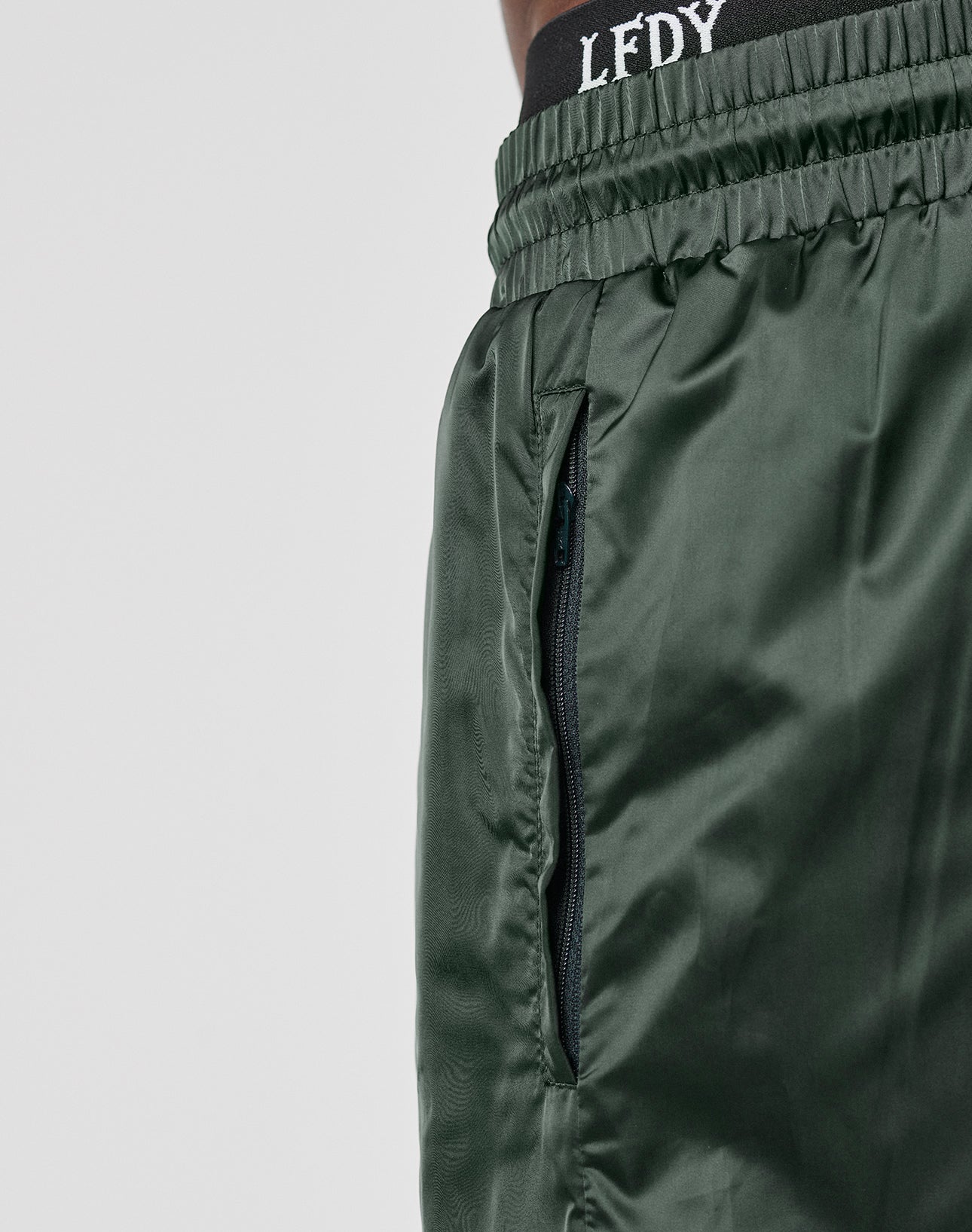 Close-up of a person wearing LIVE FAST DIE YOUNG Basic Satin Shorts—shiny, dark green polyester with elastic waistband, zippered pocket, and white textured “LFDY” letters. Regular fit for comfort and style.