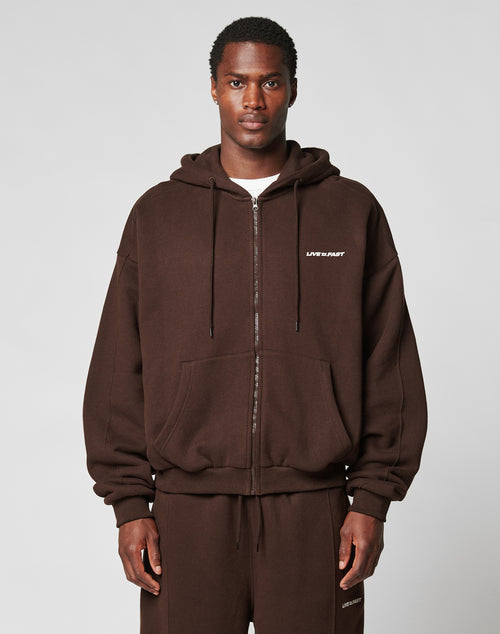 A man wears an oversize fit, dark brown LFDY Pleated Hooded Zip by LIVE FAST DIE YOUNG with LIFE IS PART text on the left chest and matching sweatpants, standing against a plain light gray background.