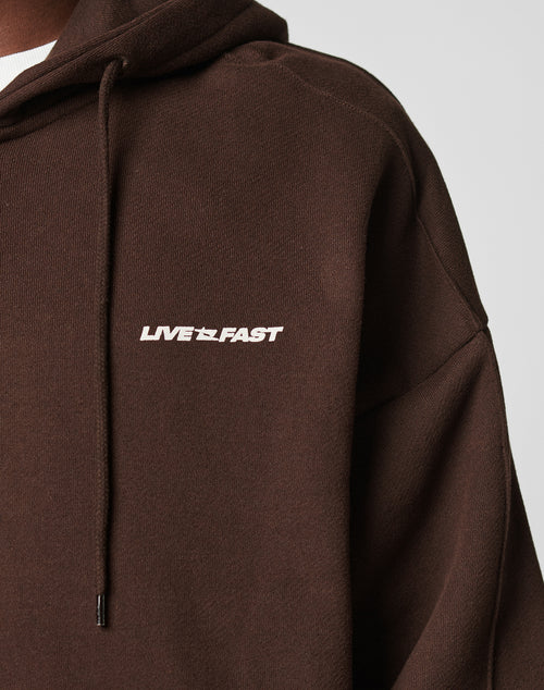 A close-up of a person wearing the LFDY Pleated Hooded Zip by LIVE FAST DIE YOUNG, an oversized brown hoodie with LIVE FAST in white on the chest, featuring a Kapuze mit Kordel and crafted from 100% Baumwolle for comfort.