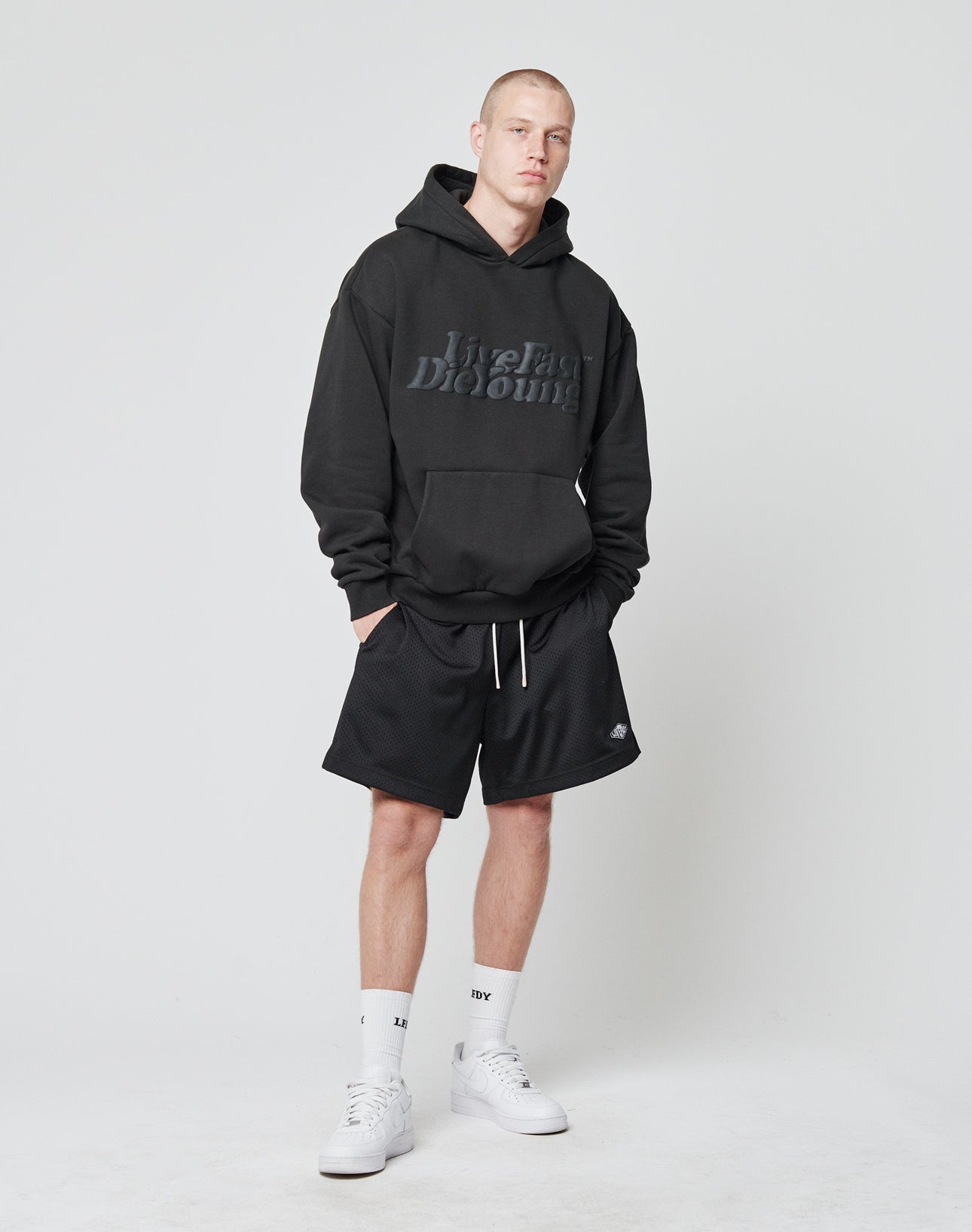 A man stands against a plain background, wearing the LIVE FAST DIE YOUNG Basic Hooded in black, paired with black shorts, white crew socks, and white sneakers. He faces the camera neutrally with hands in his hoodie pocket.