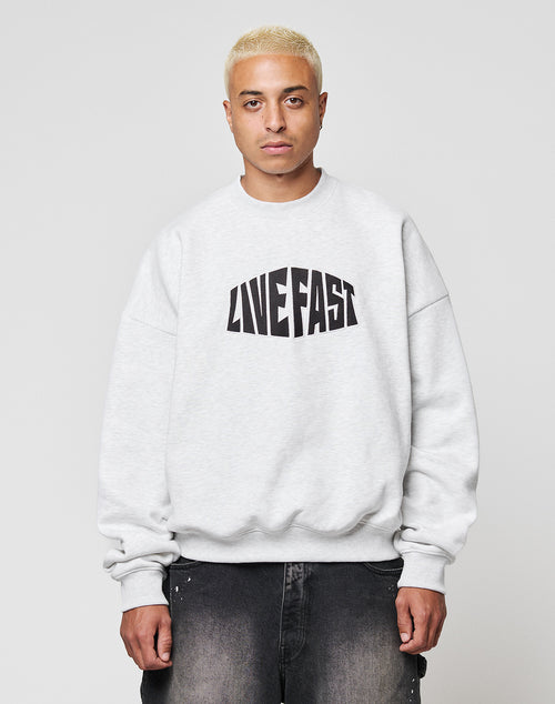 A person with short blond hair wears the LFDY Emblem Sweater by LIVE FAST DIE YOUNG, an oversized light gray cotton sweatshirt with bold black LIVE FAST lettering, paired with dark jeans against a plain light background.