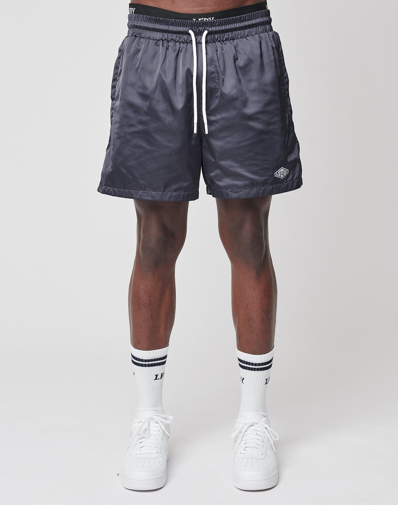 Wearing LIVE FAST DIE YOUNG Basic Satin Shorts in dark gray, paired with white crew socks featuring black stripes and white sneakers, a person stands against a plain light background. Only the lower half of their regular fit is visible.