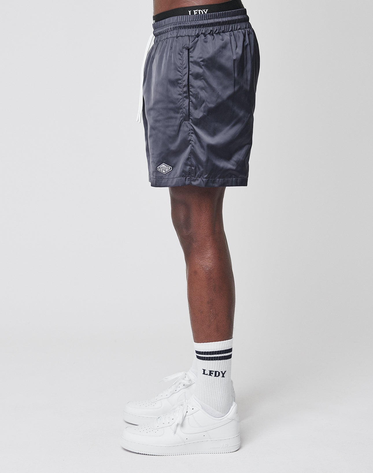 A person stands sideways against a plain white background wearing LIVE FAST DIE YOUNG Basic Satin Shorts in dark gray, paired with white socks featuring black stripes and LFDY text, and white sneakers.