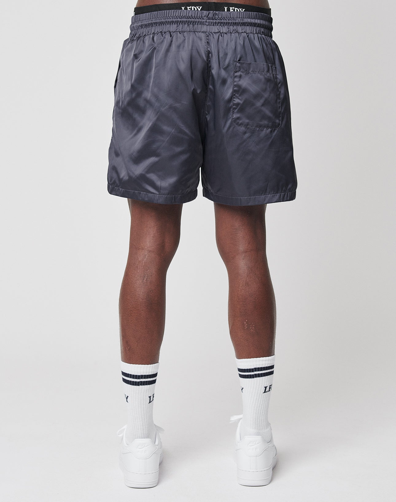 A person is seen from behind wearing LIVE FAST DIE YOUNG Basic Satin Shorts in shiny dark gray, paired with white socks featuring black stripes and white sneakers, set against a plain light-colored background.