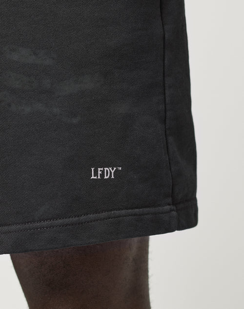 A close-up shows a person wearing the LIVE FAST DIE YOUNG Basic Sweat Shorts in black with an oversized fit and a white LFDY™ logo embroidered near the hem, set against a plain, minimal background.