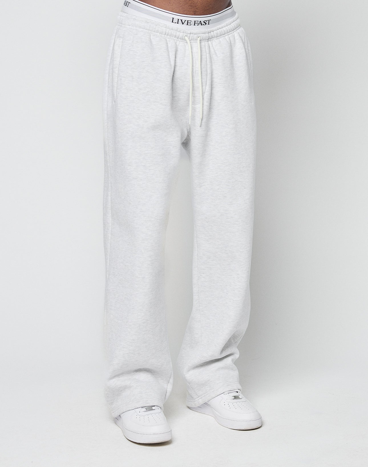A person wearing LIVE FAST DIE YOUNG Basic Wide Jogger sweatpants in light gray with an elastic waistband and drawstring, white sneakers, and underwear with LIVE FAST printed on the elastic band, standing before a plain white background.