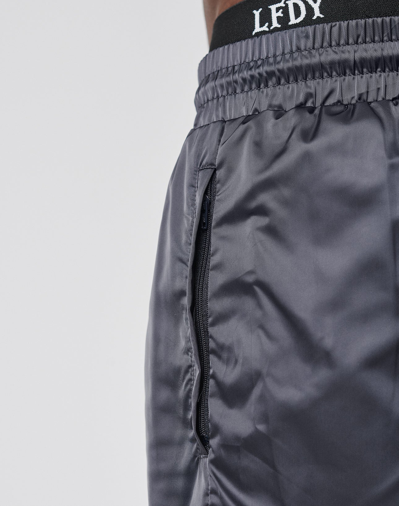 Close-up of a person wearing LIVE FAST DIE YOUNG Basic Satin Shorts—shiny gray polyester with a zippered side pocket and white LFDY text on a black waistband. A subtle logo patch accents the regular fit. Plain, light-colored background.
