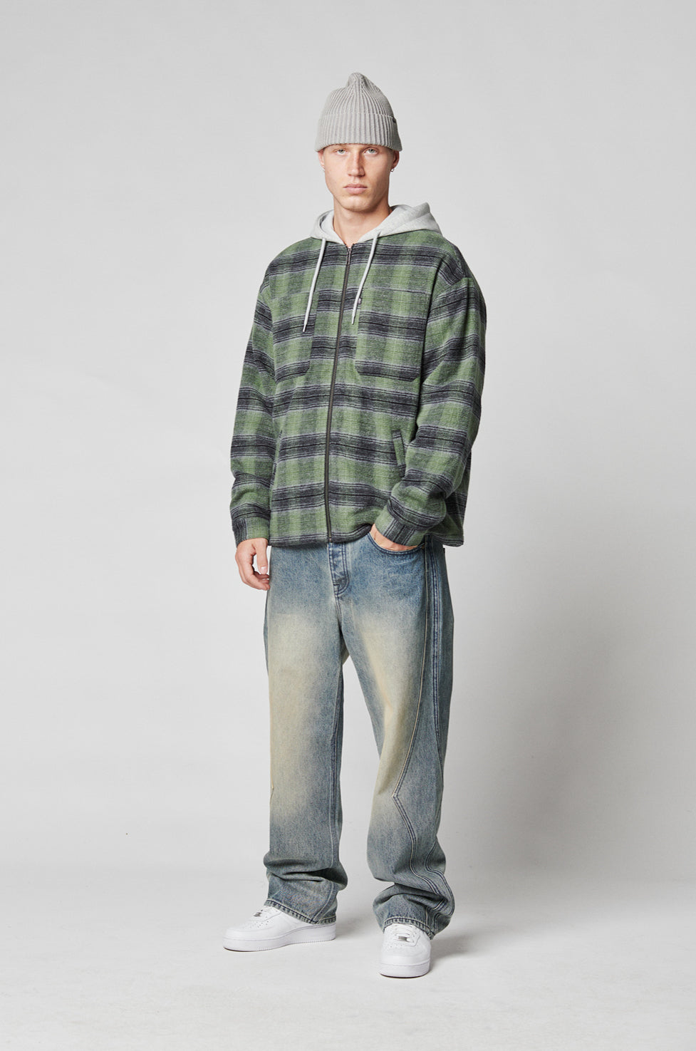 A man wears LIVE FAST DIE YOUNGs Rhomb Wide Denim jeans with a gray beanie, green plaid zip-up jacket over a hoodie, and white sneakers, standing against a plain background and looking at the camera neutrally.
